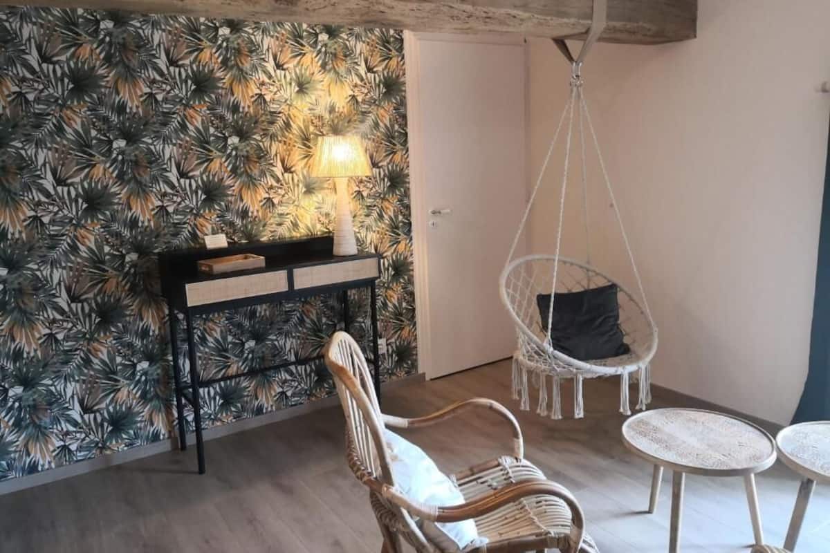 A room with a tropical leaf-pattern wallpaper, a wicker chair, a hanging chair, and a small table.