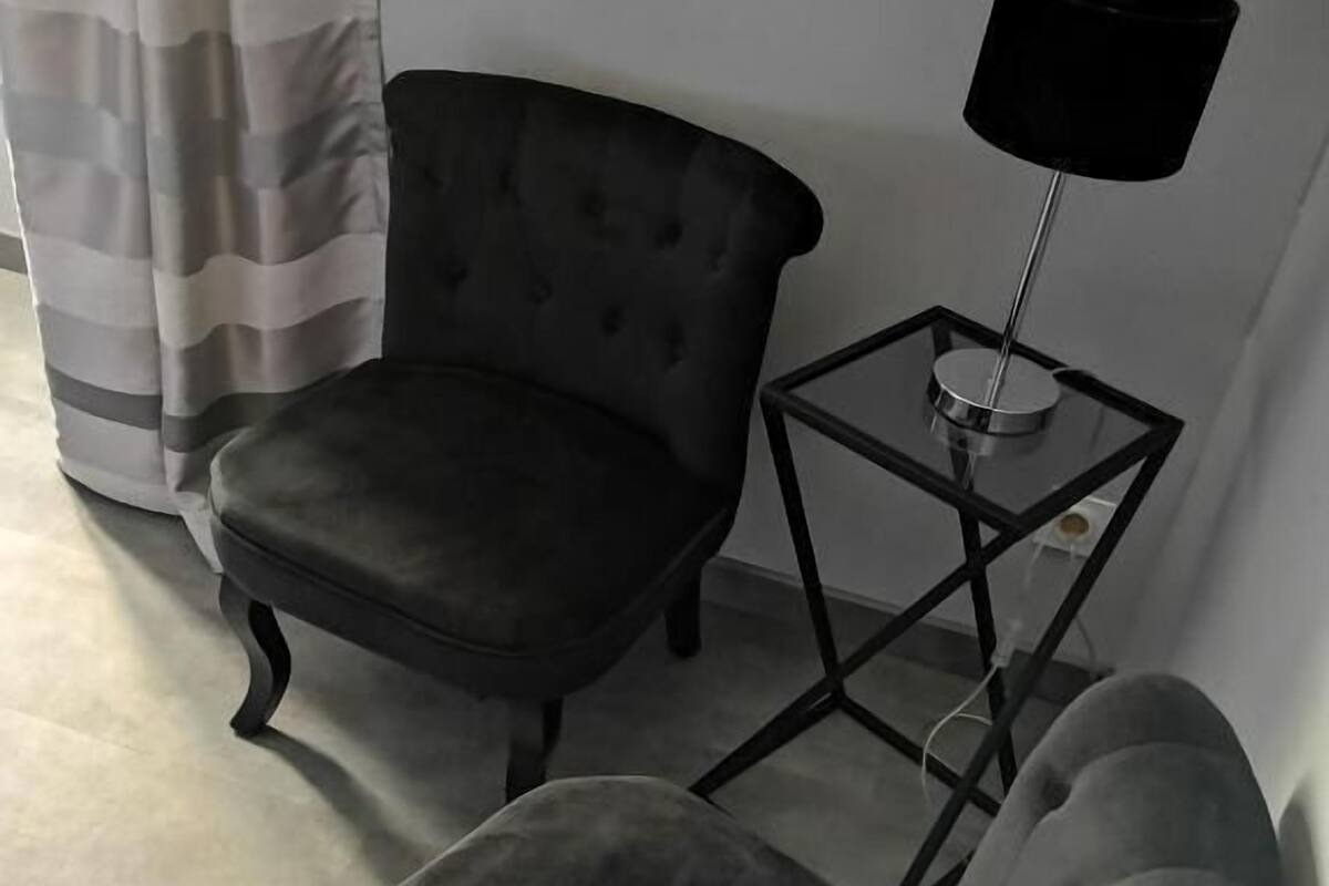 A room with a dark upholstered armchair, a smaller armchair, and a table with a lamp.