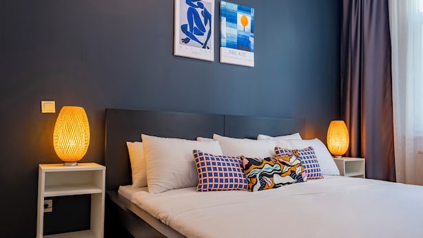 A modern hotel room with a large bed, white bedding, and colorful pillows. There are two wall-mounted artworks and two bedside lamps. - Spacious 3-Bedroom, 2-Bathroom Family Apartment in Central Location No Stair (İstanbul)