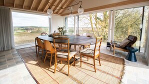 Een eetruimte met een grote ronde tafel, houten stoelen en uitzicht op het landschap door grote ramen.