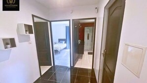 Ein Hotelgang mit schwarzem Fliesenboden, ein Bett, das durch eine geöffnete Tür erkennbar ist, und eine kleine Ablagefläche mit einem Topfpflanzen.