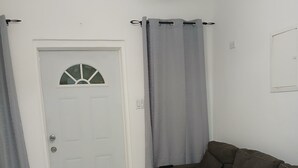 Een woonkamer met een witte deur, grijze gordijnen, een bruine bank en een bijpassend pouf.
