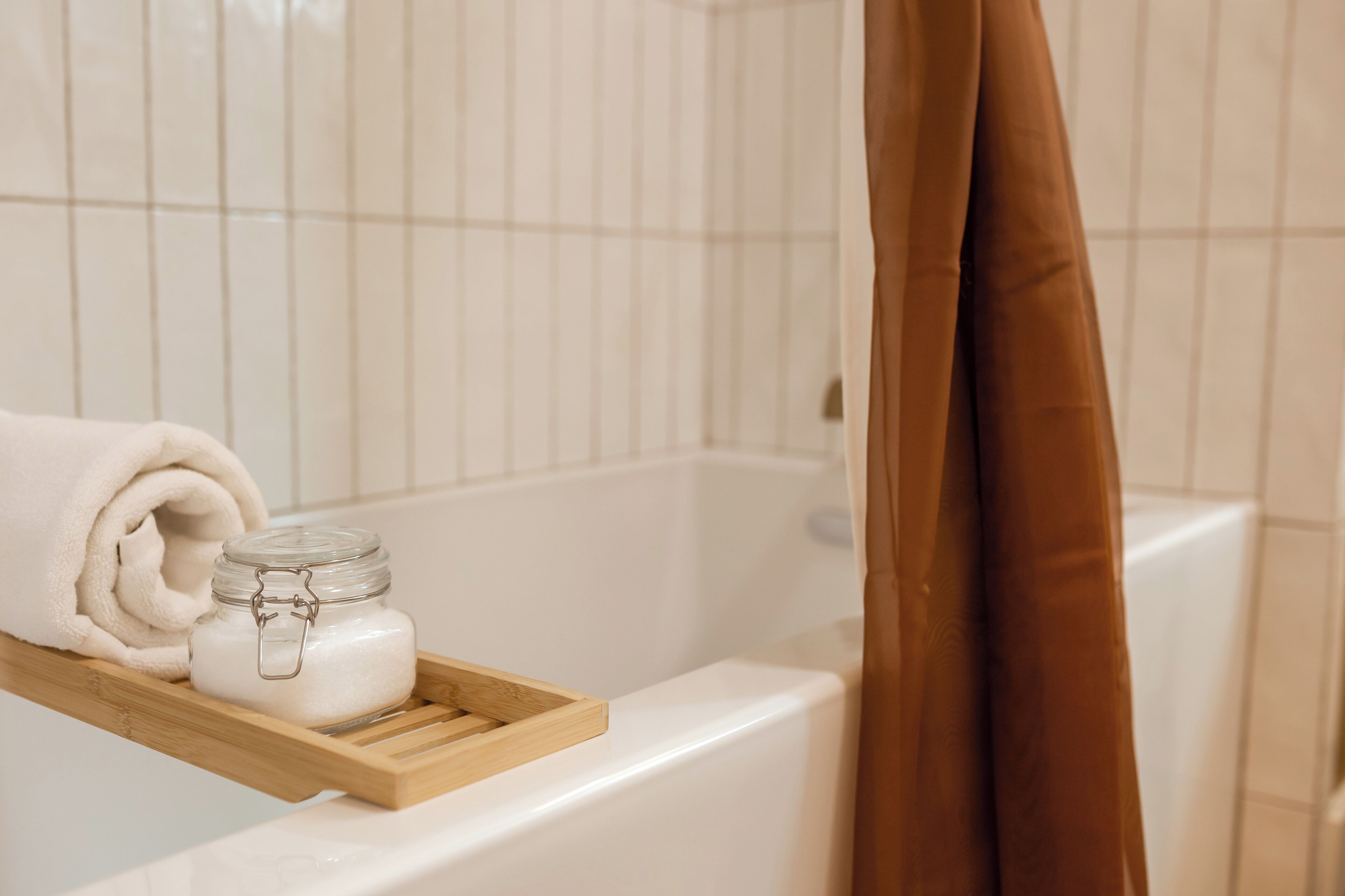 A bathroom with a white bathtub, a wooden tray holding a jar of white cream, and a rolled white towel.
