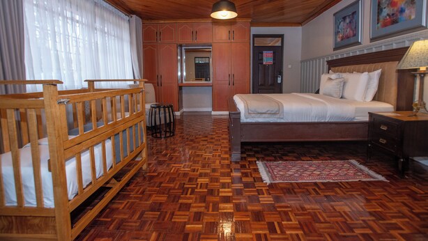 A wooden-floored bedroom with a bed, a wooden crib, and a small kitchen area. - King Suite with Balcony and Bathtub near UN Nairobi, Cozy Stead Garden House (Nairobi)