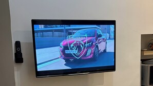Seinälle asennettu litteä näyttö, jossa näkyy auto, puinen pöytä, jossa on kynttilä, ja lasipurillinen korkkeja.