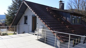 Een modern huis met een schuin dak, een opvallende schoorsteen en een balkon met een metalen hekwerk.