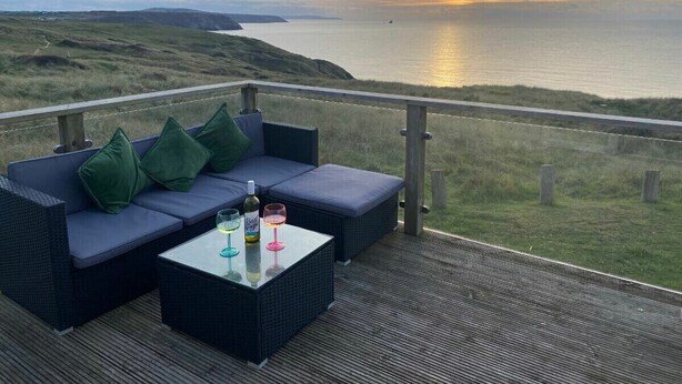 Ein Balkon mit einer Couch und einem Tisch mit Blick auf eine Küstenlandschaft bei Sonnenuntergang.