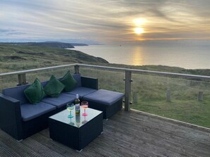 A balcony with a sofa and table, overlooking a coastal landscape during sunset. - Perran Sands - Holiday Accommodation 21444 (Perranporth)