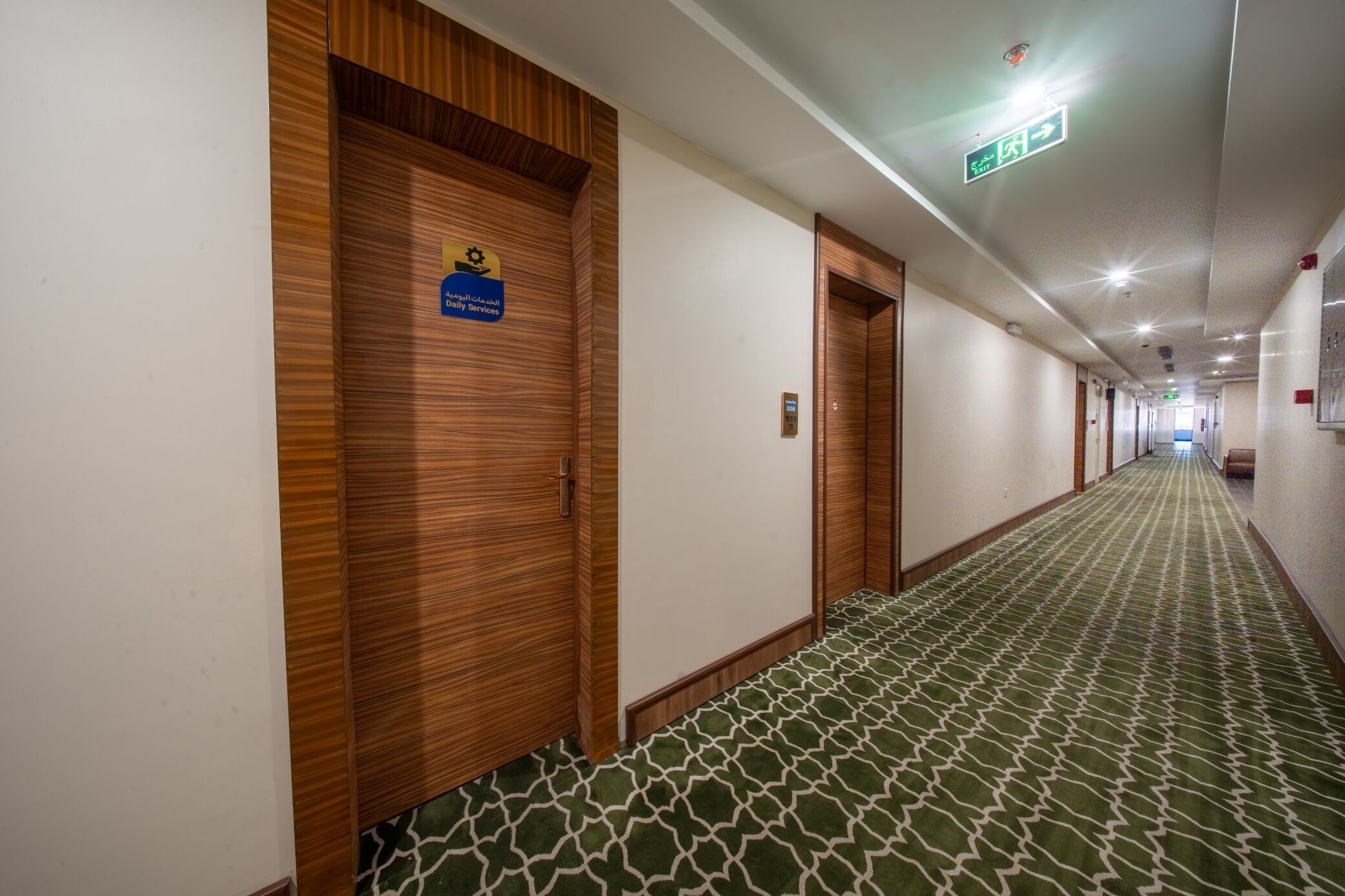 A hotel corridor with wooden doors, a patterned carpet, and a sign indicating 'Royal Saudi Daily Services.'