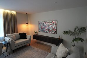 Ein modernes Wohnzimmer mit grauem Sofa, einem schwarzen Beistelltisch, einem grauen Sessel, einem gemusterten Teppich, einem dunklen Holzkonsolentisch, einem an der Wand befestigten abstrakten Kunstwerk und einer Topfpflanze.