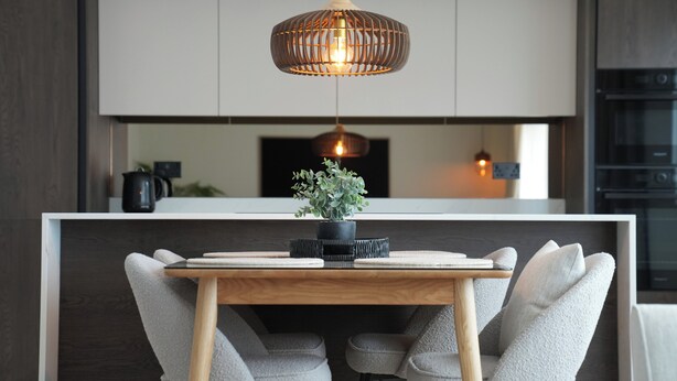A modern kitchen with a central island, two grey upholstered chairs, a wooden table, and a hanging pendant light. - 1st Floor Luxury 2-bed Apt, Sleeps 6 (St. Andrews)