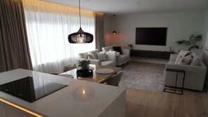 A modern living room with a large flat-screen TV, a dining area with a white table and chairs, and a kitchen with a built-in stove. - 1st Floor Luxury 2-bed Apt, Sleeps 6 (St. Andrews)