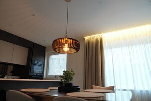 Uma área de jantar moderna com uma mesa central, cadeiras, um lustre pendurado, uma planta em vaso e uma janela com cortinas.