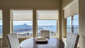 A dining area with a round table and four striped chairs, large windows with a view of a distant landscape and water. - Heavenly Pointe | Penthouse Lake Views, Elevator, Accessible, King Beds (Branson)