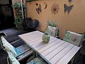 Um pátio externo com mesa de madeira, cadeiras com padrão verde e azul, um vaso decorativo e arte nas paredes, incluindo um sol e uma lua crescente.