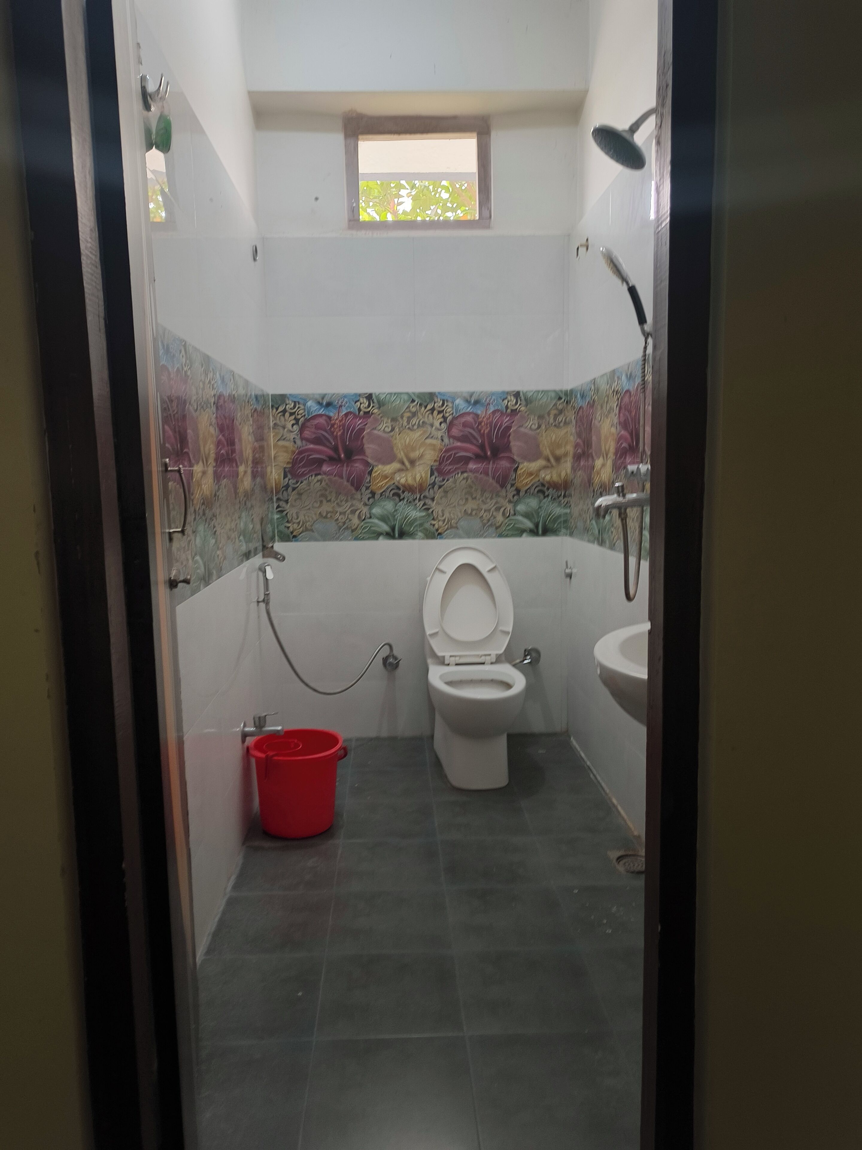 A small bathroom with a toilet, a sink, and a shower. There is a window above the toilet and a red bucket on the floor.