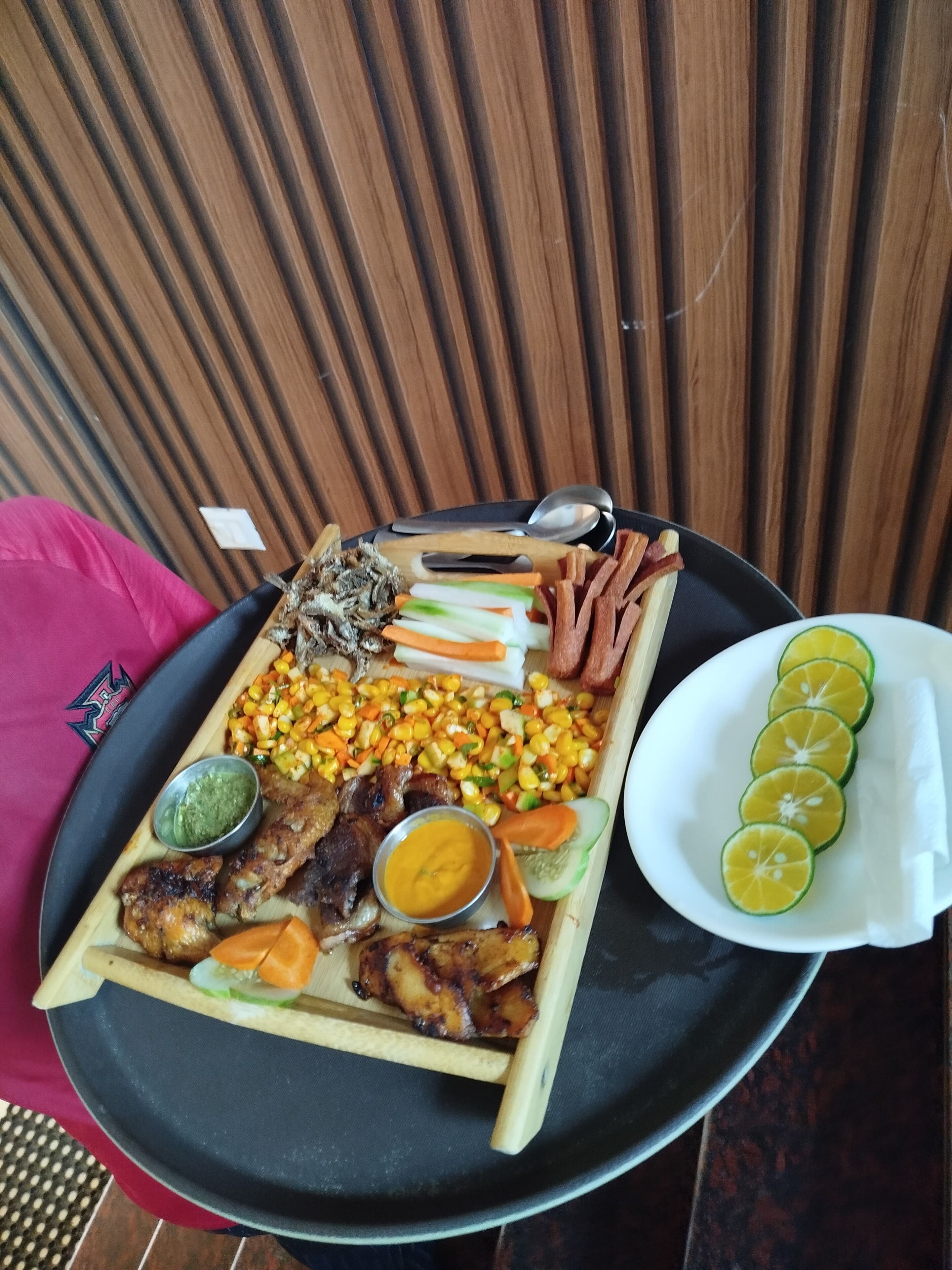 A tray of food including grilled meat, corn, carrots, and lime slices.
