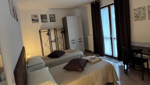 Une chambre d’hôtel avec deux lits, un bureau et une chaise. On y trouve un radiateur, une fenêtre avec des rideaux et des œuvres d’art accrochées aux murs.