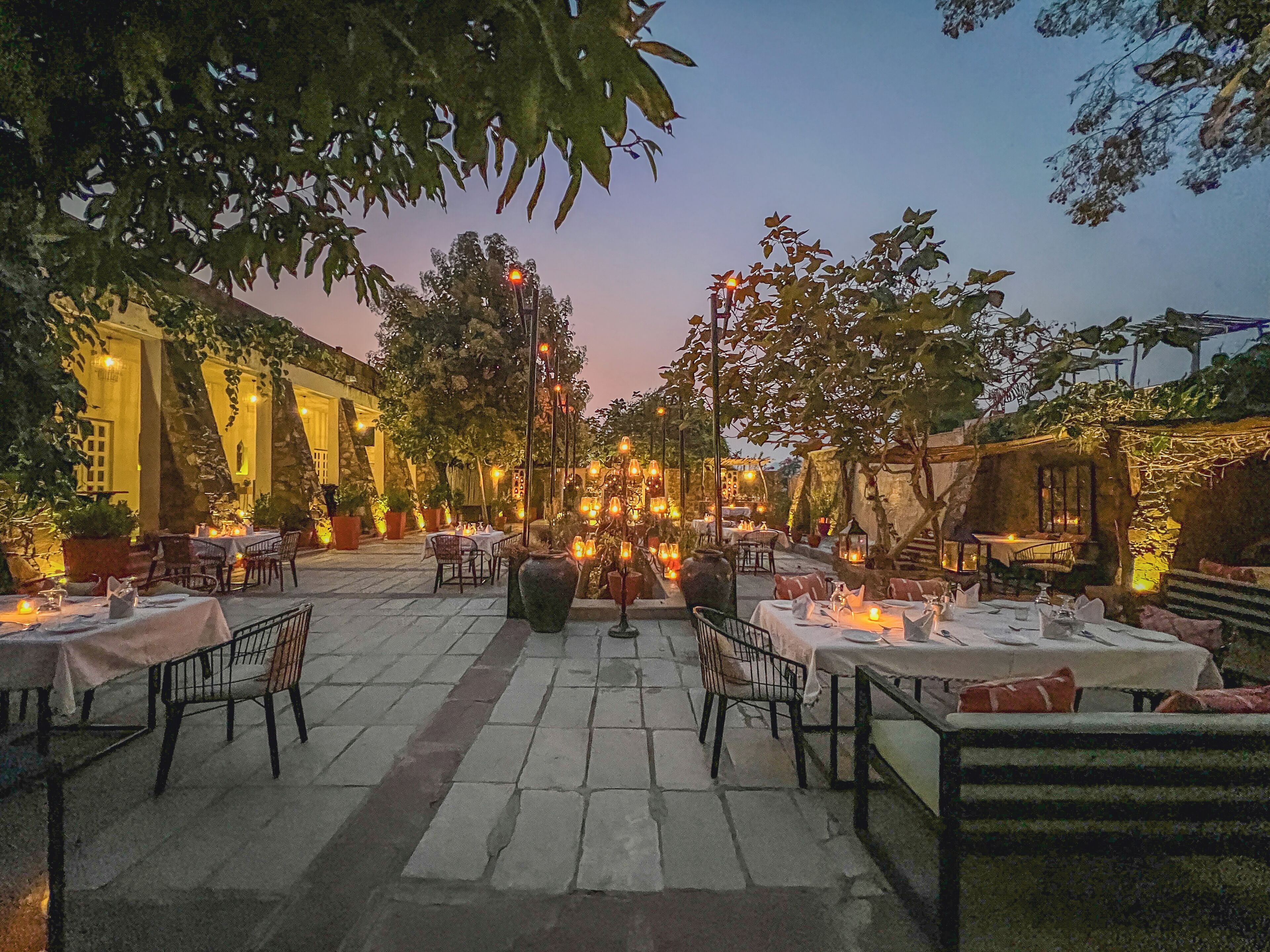 An outdoor dining area with tables and chairs, surrounded by trees and stone flooring.