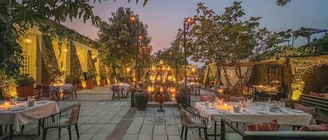An outdoor dining area with tables and chairs, surrounded by trees and stone flooring.