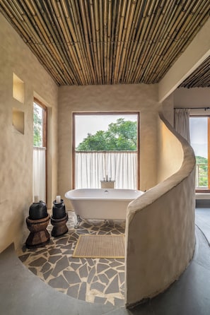 A bathroom with a freestanding bathtub, a window with a view of greenery, and a curved wall. - Utsav Camp Sariska (Rajgarh)