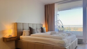 A modern bedroom with a large bed, a bedside table with a lamp, and a view of a balcony with a chair. - Ocean Suite von OstseeAhoi (Timmendorfer Strand)