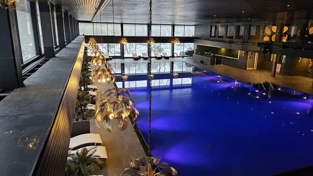A modern indoor swimming pool with a long, narrow seating area along one side, large windows, and hanging pendant lights. - Ocean Suite von OstseeAhoi (Timmendorfer Strand)