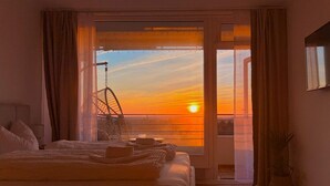 A bedroom with a bed, a chair, a plant, and a view of the sunset through a window. - Ocean Suite von OstseeAhoi (Timmendorfer Strand)