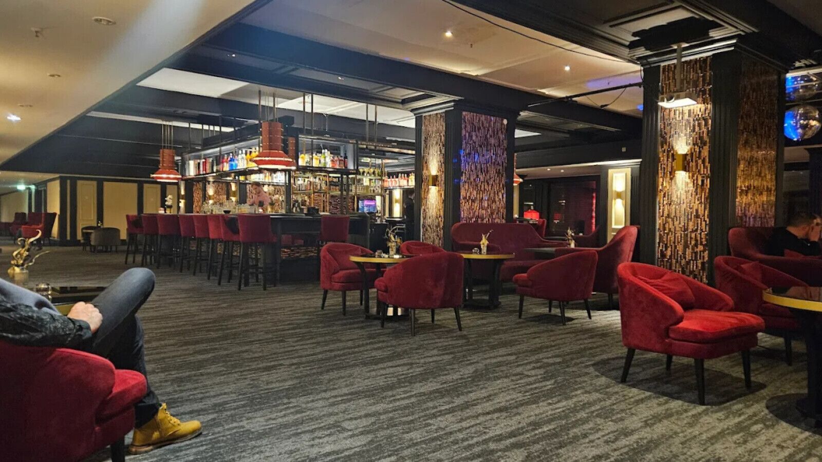 Um hall de hotel com cadeiras estofadas vermelhas, área de bar com prateleiras de garrafas e parede com padrão.