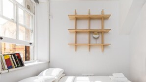 Un dormitorio minimalista con cama, una ventana llena de libros y un estante de madera con un espejo redondo.