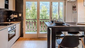 Una cocina moderna con techo de madera, un comedor con mesa y sillas, y amplios ventanales correderos que dan a una terraza con vistas a árboles.