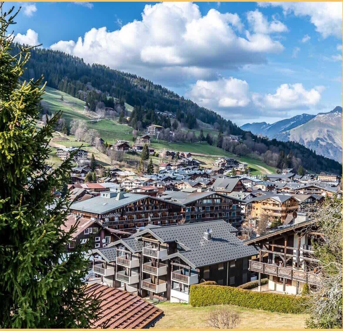 A mountain resort with chalet-style buildings, a ski lift, and a view of the surrounding hills and mountains.