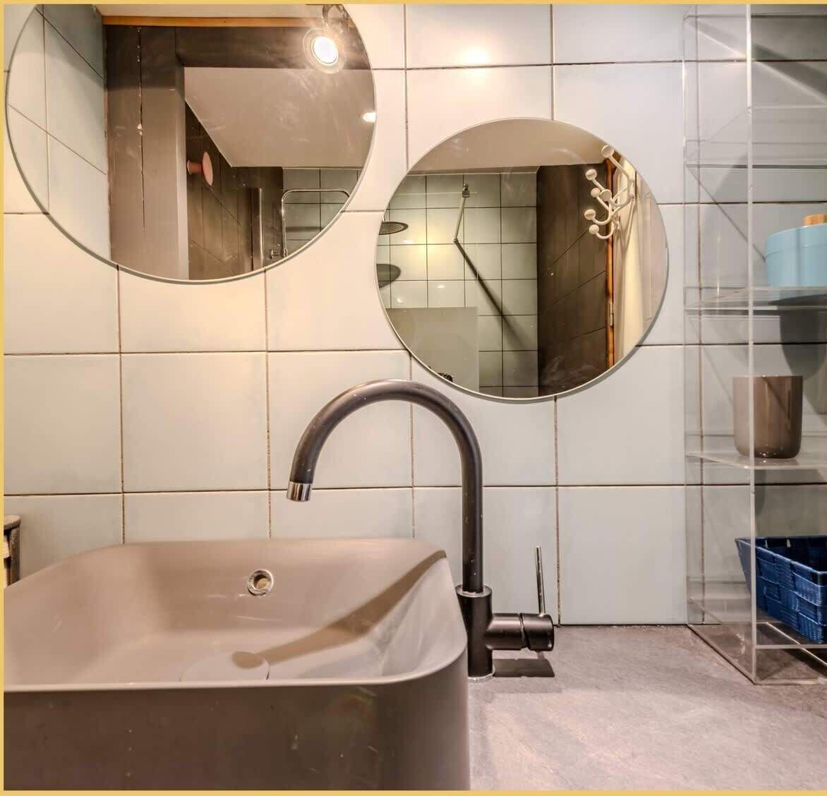 A modern bathroom with a freestanding bathtub, two wall-mounted mirrors, and a glass shelving unit.