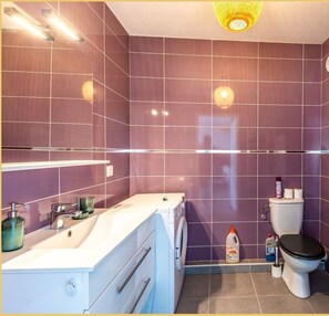 Kylpyhuoneessa on violetit laatat, valkoinen pesuallas, wc-istuin ja pesukone.