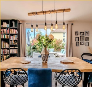 Un coin repas avec une table en bois, des chaises à l’assise bleue, un vase rempli de fleurs et une bibliothèque remplie de livres.