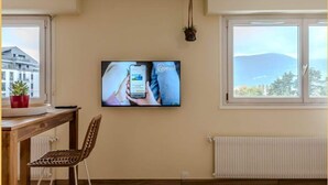 A room with a wooden floor, a dining table with a chair, a TV mounted on the wall, and a window with a view of a mountain. - Chez Carole I Studio I Annemasse (Annemasse)