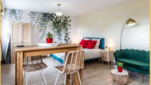 A modern living room with a wooden dining table, two wicker chairs, a green sofa, a bedside table with a lamp, and a wall-mounted plant. - Chez Carole I Studio I Annemasse (Annemasse)