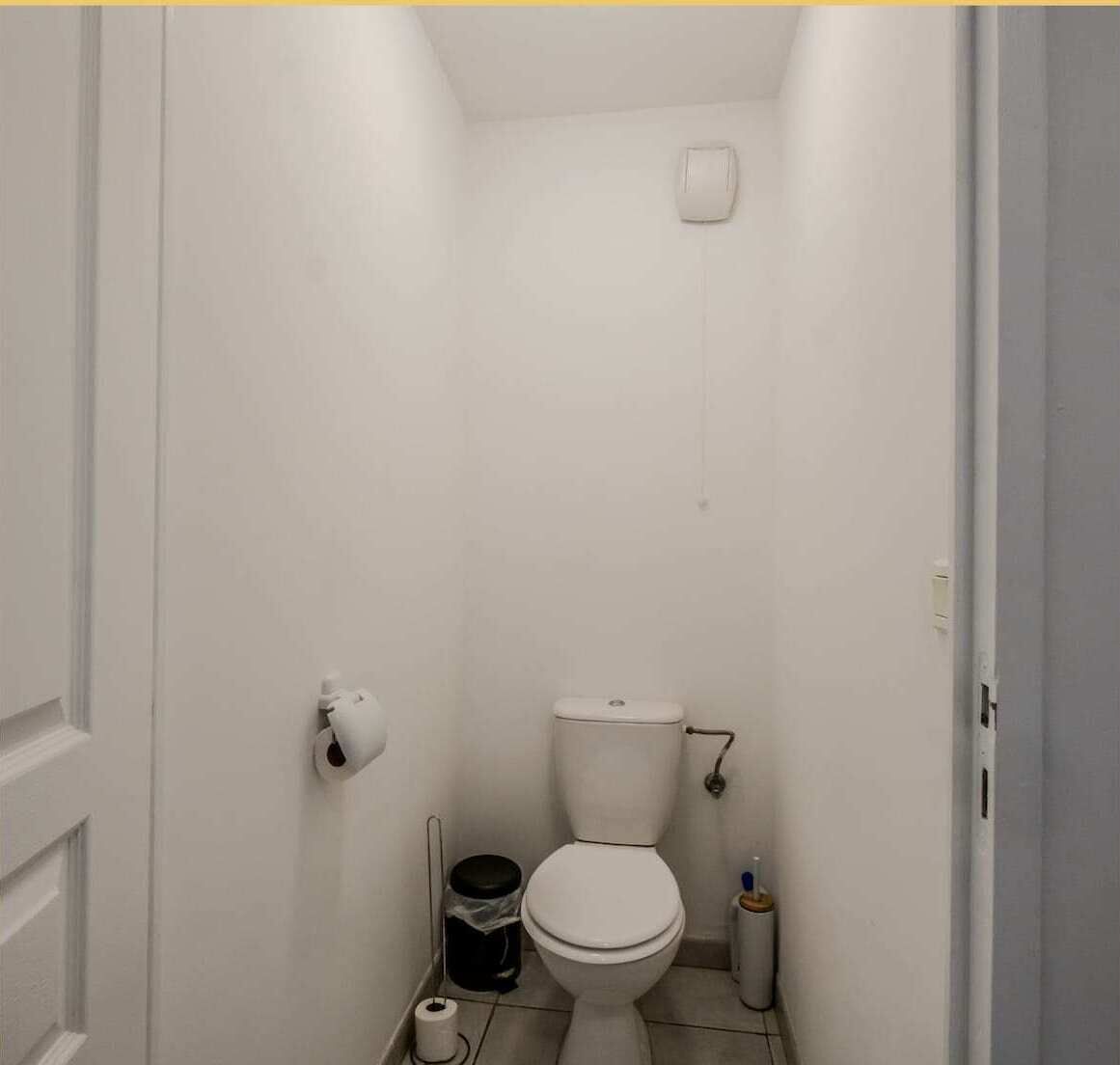 A small, narrow bathroom with white walls, a toilet, a trash bin, and a toilet paper holder.