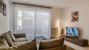 A living room with a sofa, a TV, a lamp, and sheer curtains. - Le Manoir de Jérémy I T2 I Thonon (Thonon-les-Bains)