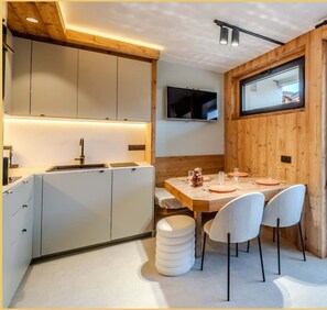 A modern kitchen with white cabinets, a wooden dining table with chairs, and a TV mounted on the wall. - L'Élégance Alpine I Centre Les Gets (Les Gets)