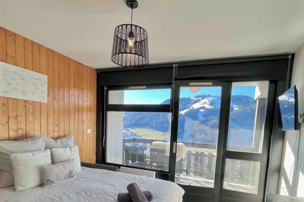 Ein Schlafzimmer mit Bett, Kissen, einer Pendelleuchte und einem großen Fenster mit Blick auf die Berge.