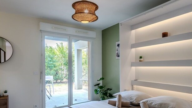 Une chambre à coucher avec un lit, un luminaire au plafond, une porte-fenêtre coulissante et une plante.