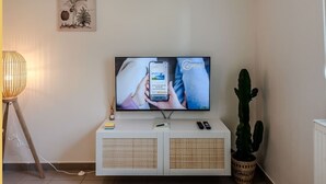 Un salon moderne avec un téléviseur à écran plat fixé à un meuble blanc, un cactus, une lampe et une illustration botanique encadrée accrochée au mur.