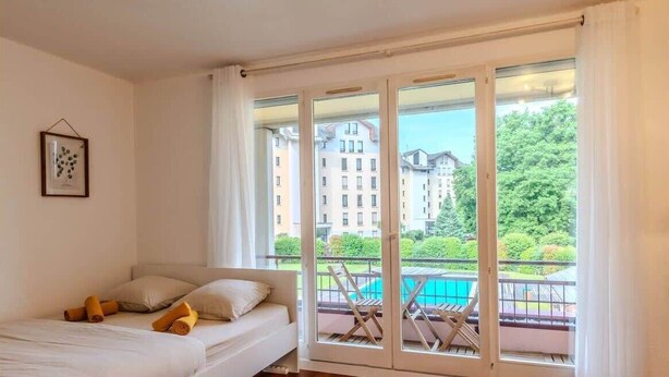 Ein Schlafzimmer mit Bett, Fenster und Blick auf einen Pool sowie Gebäude.