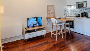 Ein kompakter Wohnraum mit Holzboden, einem Essbereich mit Tisch und Stühlen, einer Küchenzeile mit Mikrowelle und einem Fernseher auf einem weißen Schrank.