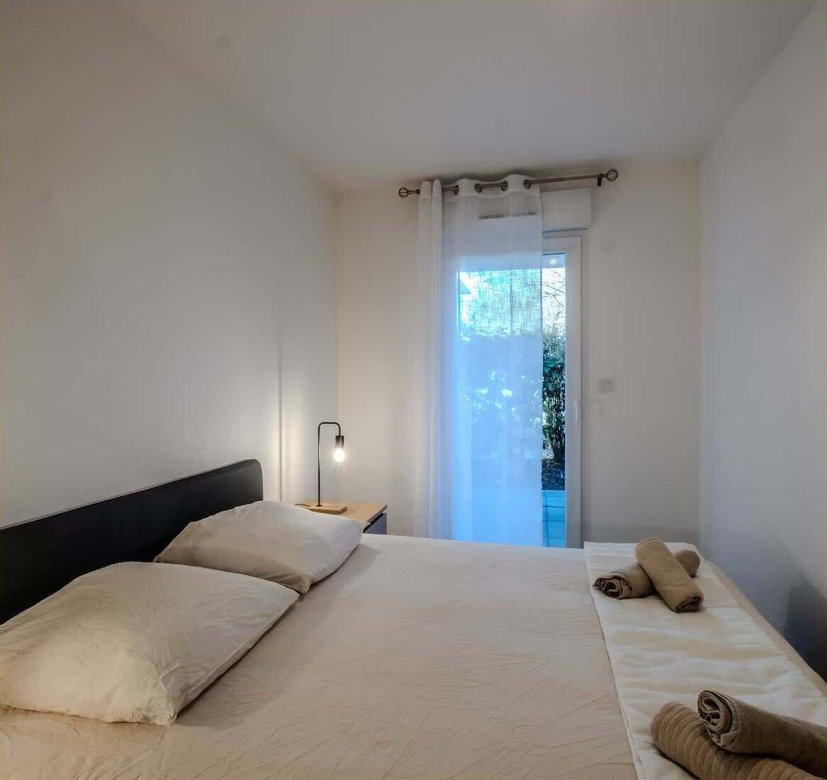 Un dormitorio con cama, mesita de noche, lámpara y vistas al exterior.