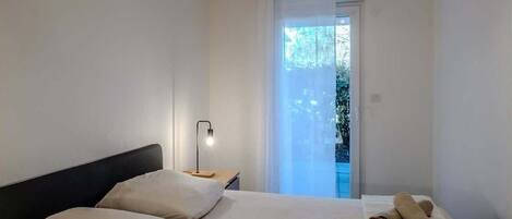 Un dormitorio con cama, mesita de noche, lámpara y vistas al exterior.