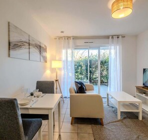 A modern living room with a dining area, a sofa, a TV, and a large window with curtains. - L'Ermitage I T2 I Thonon (Thonon-les-Bains)