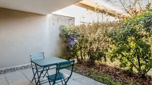 Een terras buiten met een tafel en stoelen, omgeven door groen en een gebouw op de achtergrond.