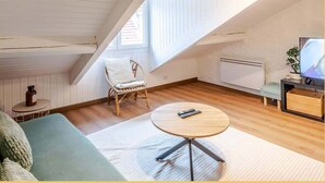A cozy attic bedroom with a sloped ceiling, a round wooden table, a wicker chair, a sofa, and a television. - Chez Noé I T2 I Thonon (Thonon-les-Bains)